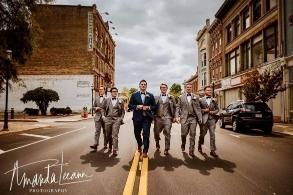 A groom and groomsmen in suits walk toward the camera down a city street.