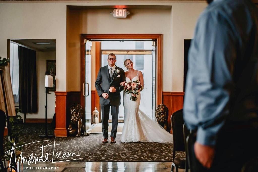 Father escorting smiling bride in a white gown down the aisle. Indoor ceremony.