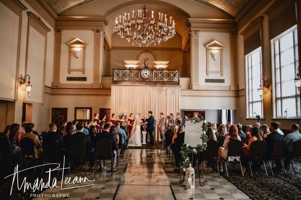 Wedding ceremony in an ornate hall. Bride and groom at the altar, guests seated. Large windows, chandelier.