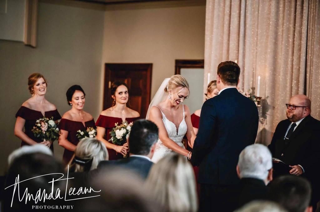 Wedding ceremony: Bride and groom exchanging rings, bridesmaids in burgundy dresses, indoors.