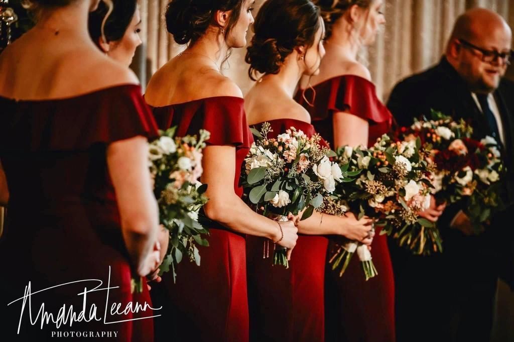 Bridesmaids in burgundy off-the-shoulder dresses holding bouquets, at a wedding ceremony.