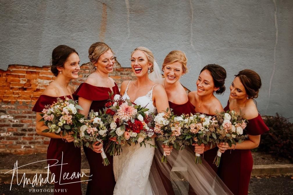 Bridesmaids in burgundy dresses laugh with the bride in front of a blue-gray wall.