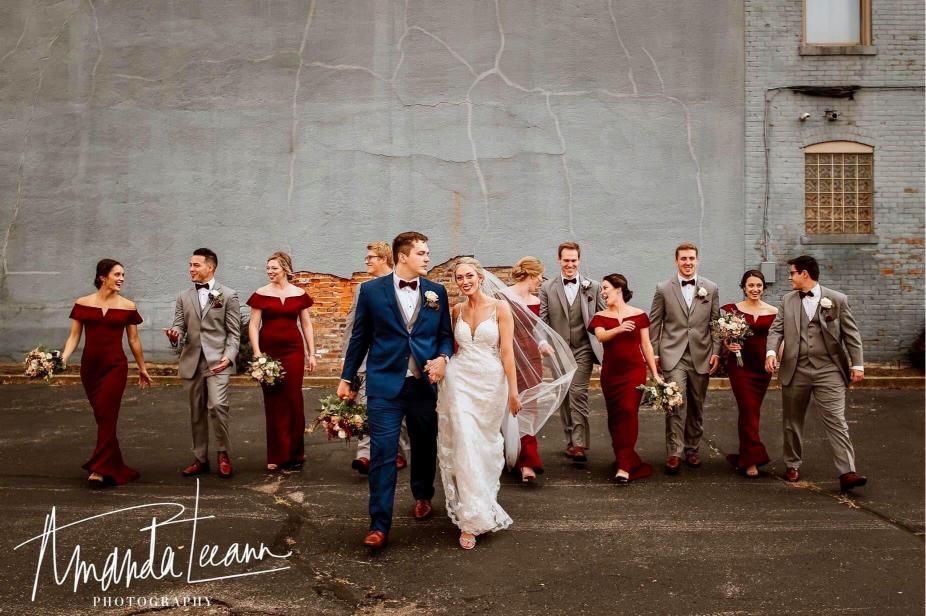 Wedding party walks toward camera; bride & groom lead, flanked by bridesmaids in red & groomsmen in gray.