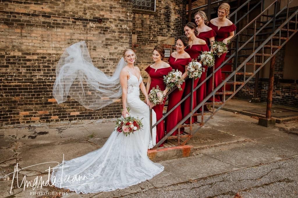 Bride in white lace gown and veil with bridesmaids in red dresses on outdoor steps.