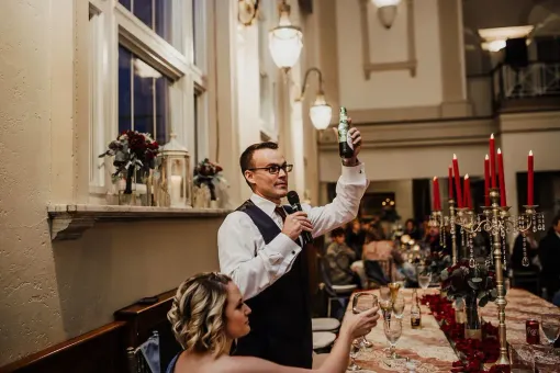 Man giving a speech at a wedding, holding up a bottle. A woman next to him raises a champagne flute.
