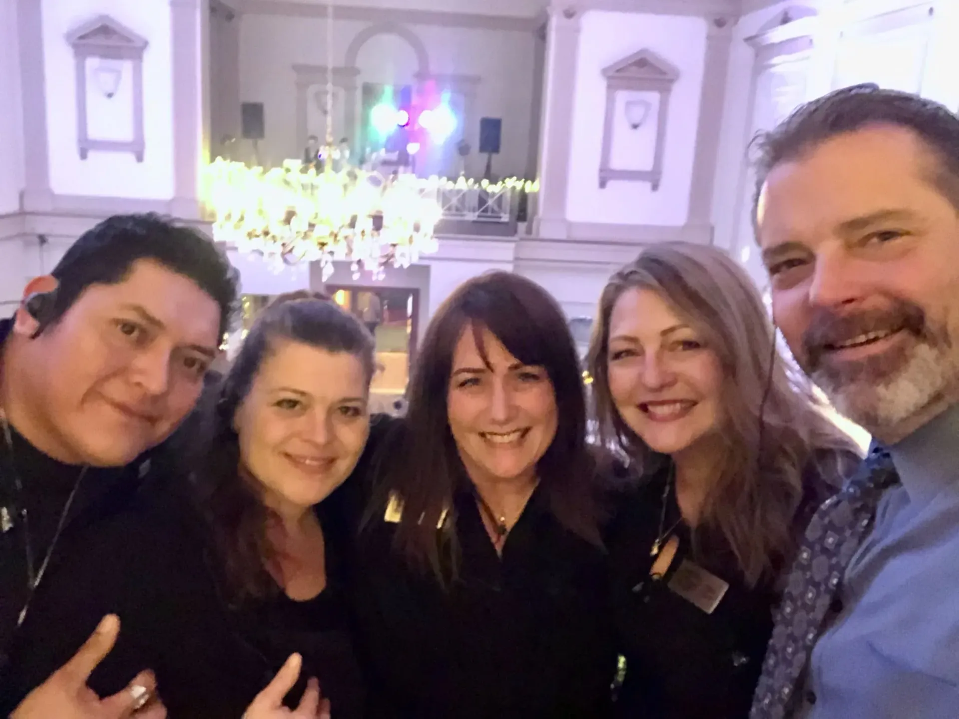 Group selfie of five people smiling inside a building with a chandelier and stage in the background.