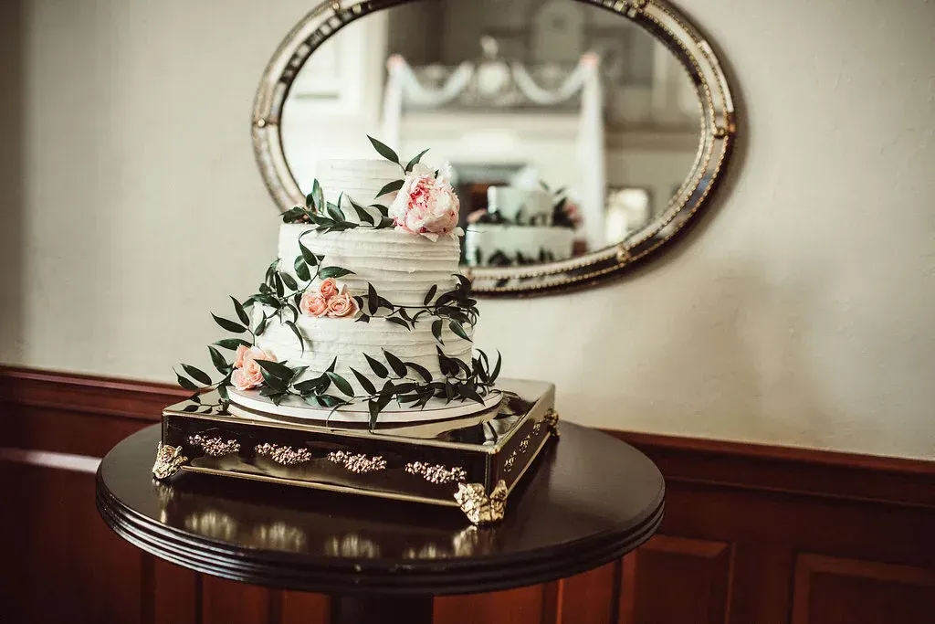 Three-tiered wedding cake decorated with flowers and greenery on a pedestal, reflected in an oval mirror on a brown table.