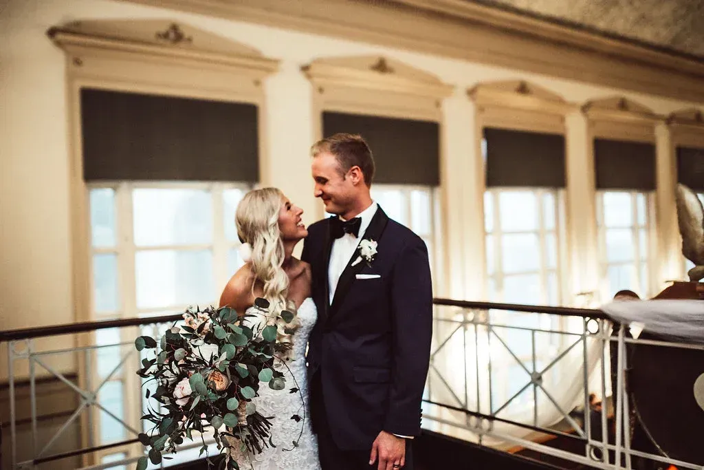 Newlyweds smiling at each other, standing on a balcony with a dark suit and a lace wedding dress.