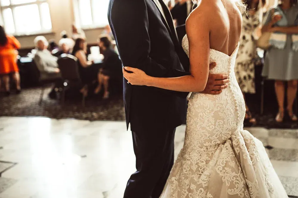 Bride and groom share a dance at their wedding reception. The bride wears a white lace gown, and the groom is in a navy suit.