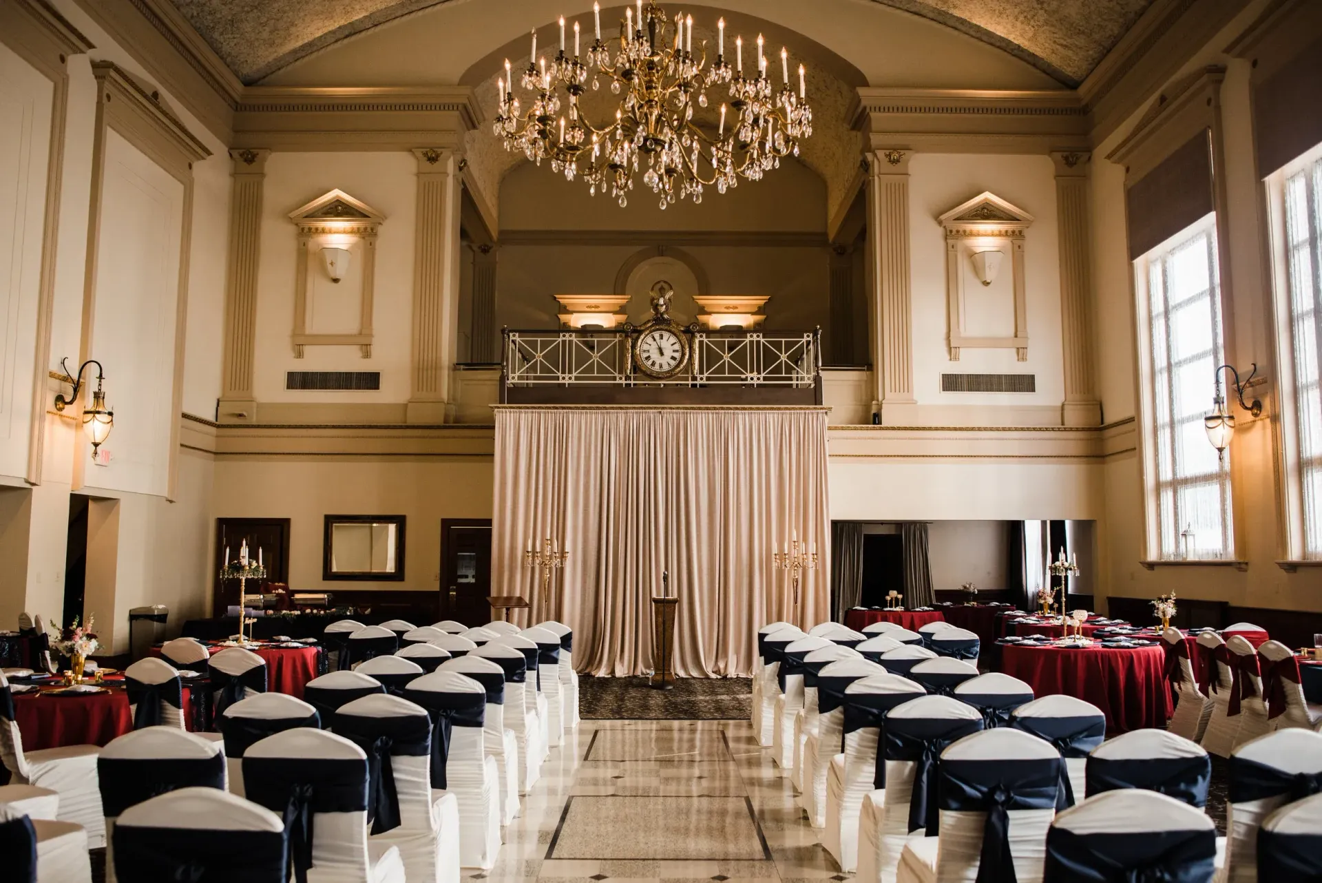 Elegant wedding ceremony setup in a grand hall with rows of chairs facing a draped stage. Gold and navy accents throughout.