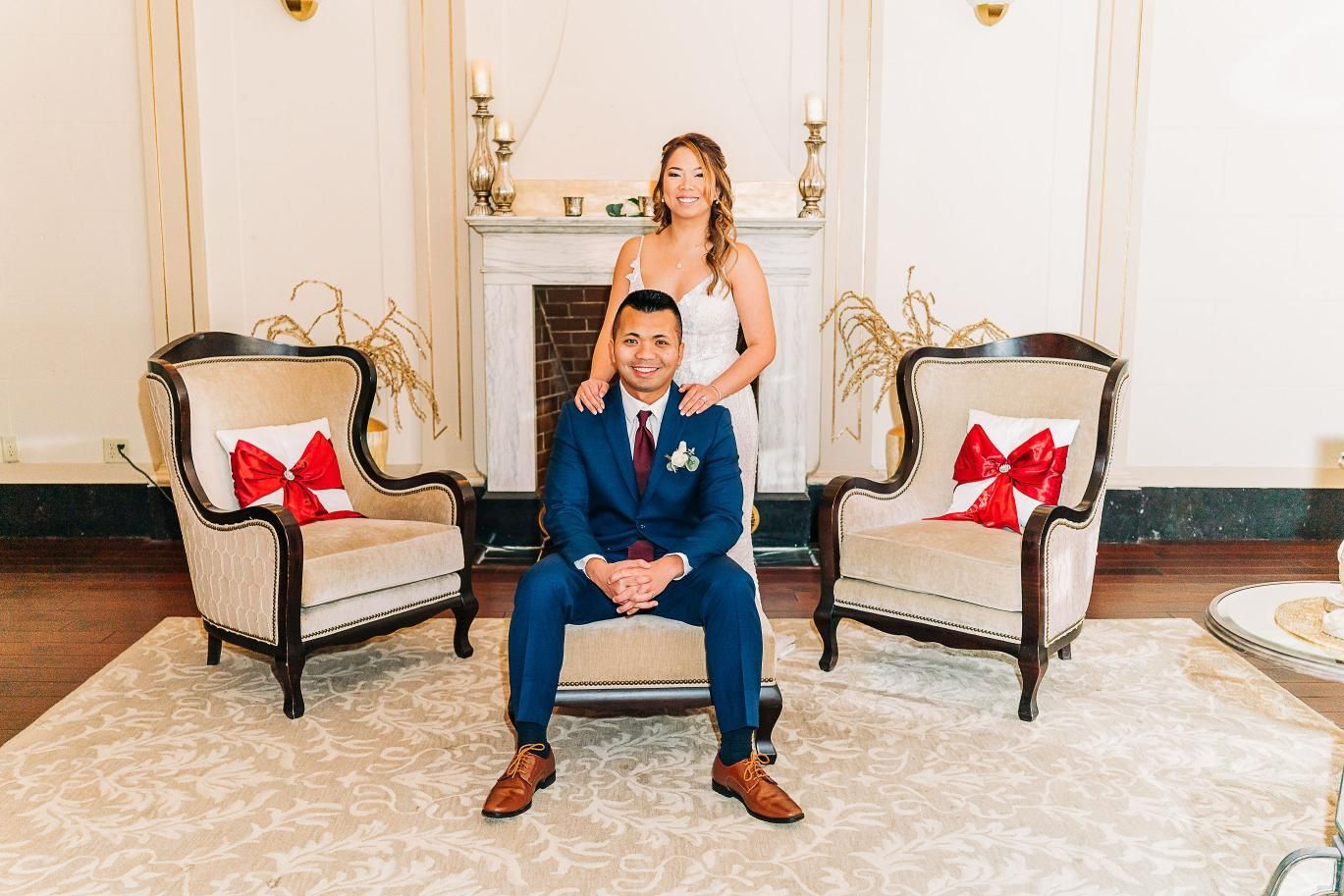 Newlyweds pose in elegant room. Man in blue suit sits in chair; woman behind him smiles.