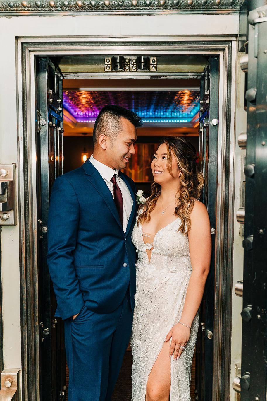 Couple in wedding attire, smiling at each other in front of a bank vault door, inside.