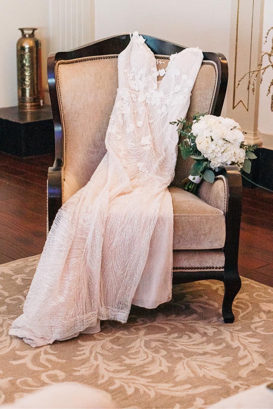 Wedding dress draped over a vintage armchair next to a bouquet of white flowers.