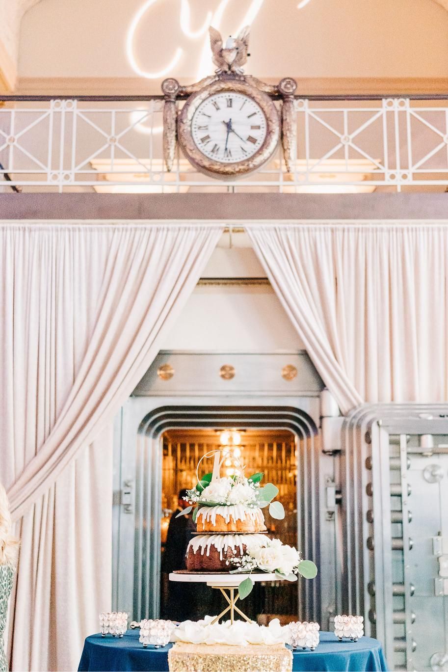 Wedding cake displayed in front of a vault door with a vintage clock and draped curtains.