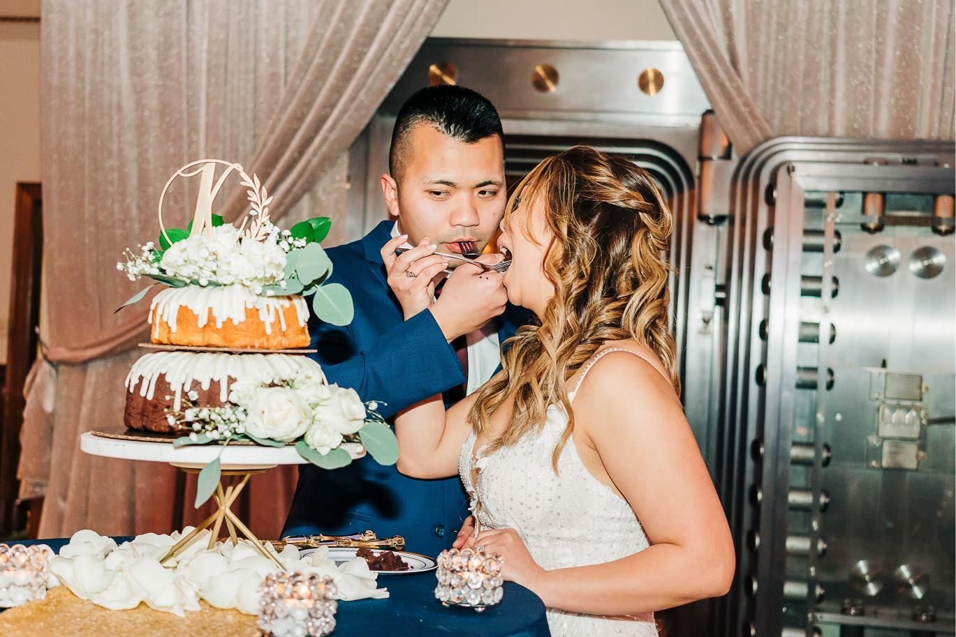 Newlyweds feed each other cake at their reception. They are in front of a vault door.