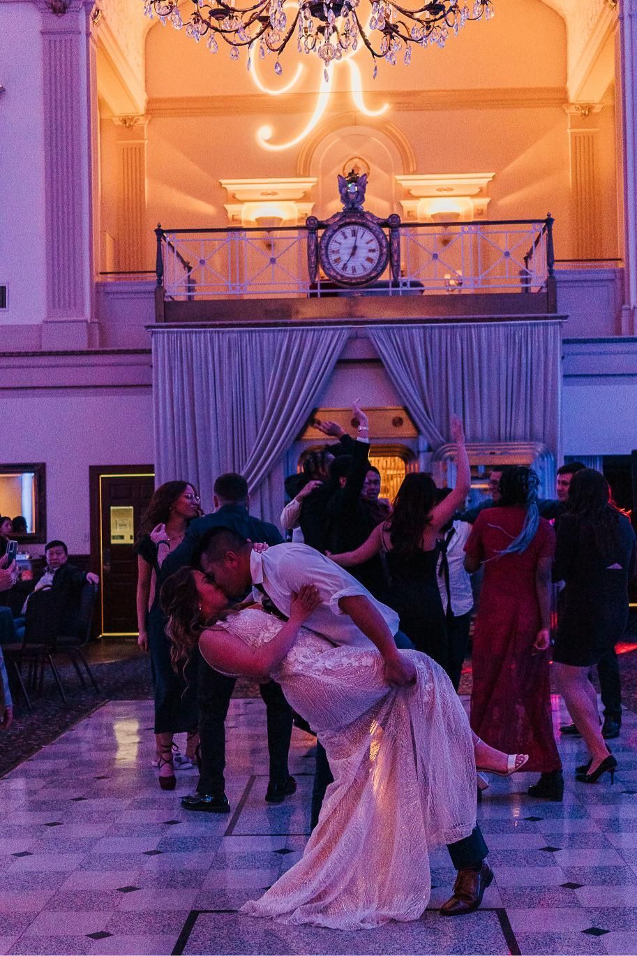 Couple dancing at a wedding reception. He dips her back. Other guests and ornate building interior in background.