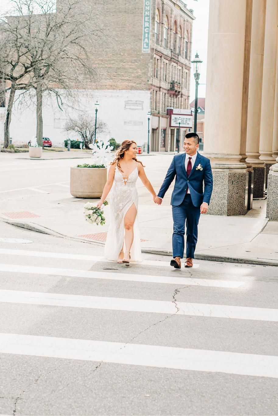 Bride and groom hold hands, walking on a crosswalk. She wears a white dress, he wears a blue suit. City setting.