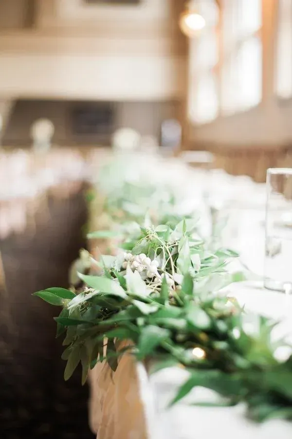 Greenery garland adorns a long table, likely for a wedding or event. Bright leaves with small white flowers.