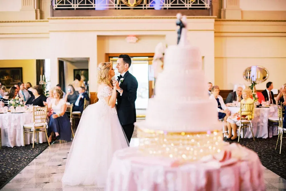 Bride and groom share a moment near a large wedding cake in a ballroom setting. Guests watch from tables.