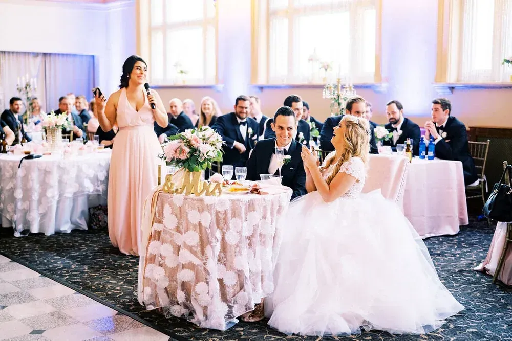 A bridesmaid gives a speech at a wedding reception. The bride and groom are at a table decorated with flowers, smiling.