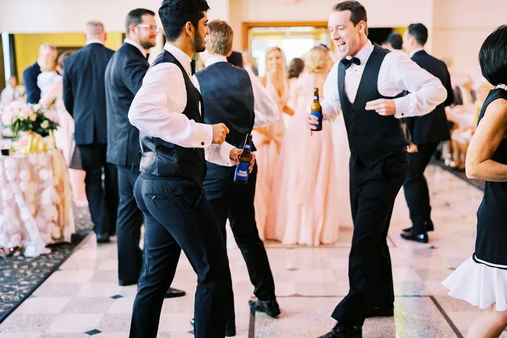Guests dancing at a wedding reception, including two men in vests and bow ties with drinks.