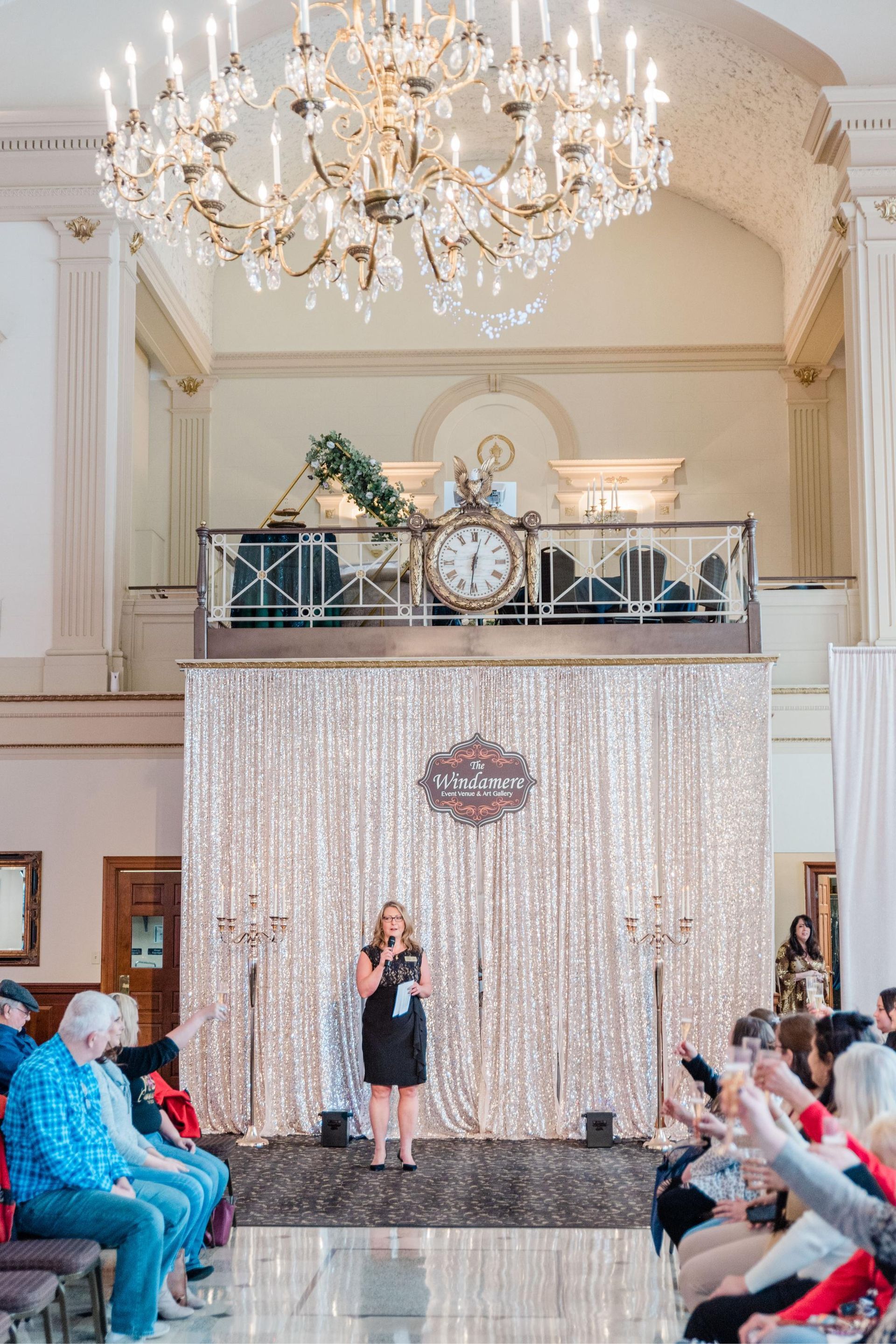 Woman speaking at a fashion event. Sparkling backdrop, chandelier, audience.