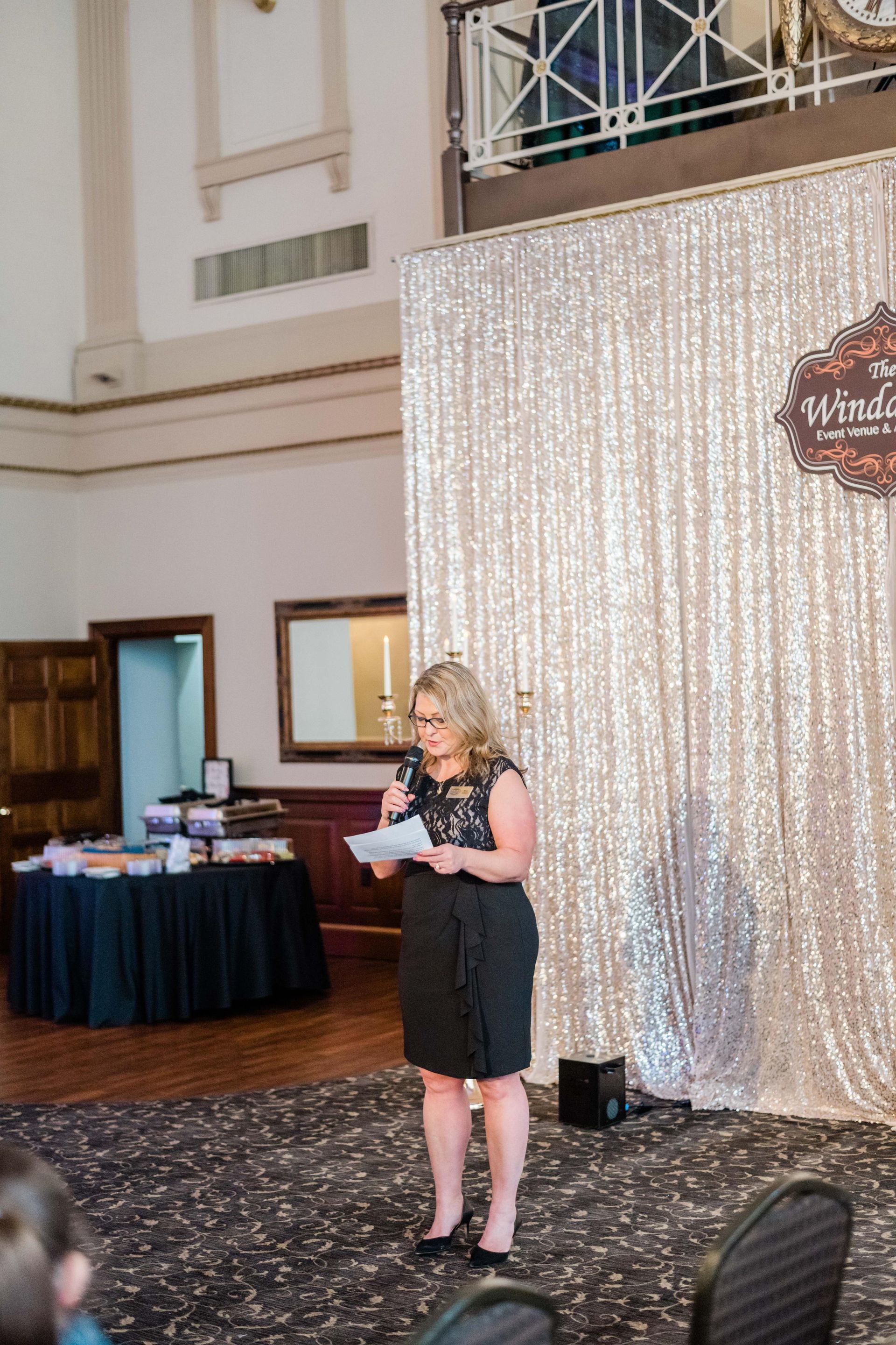 Woman in black dress speaking at a podium; sparkly backdrop; event.