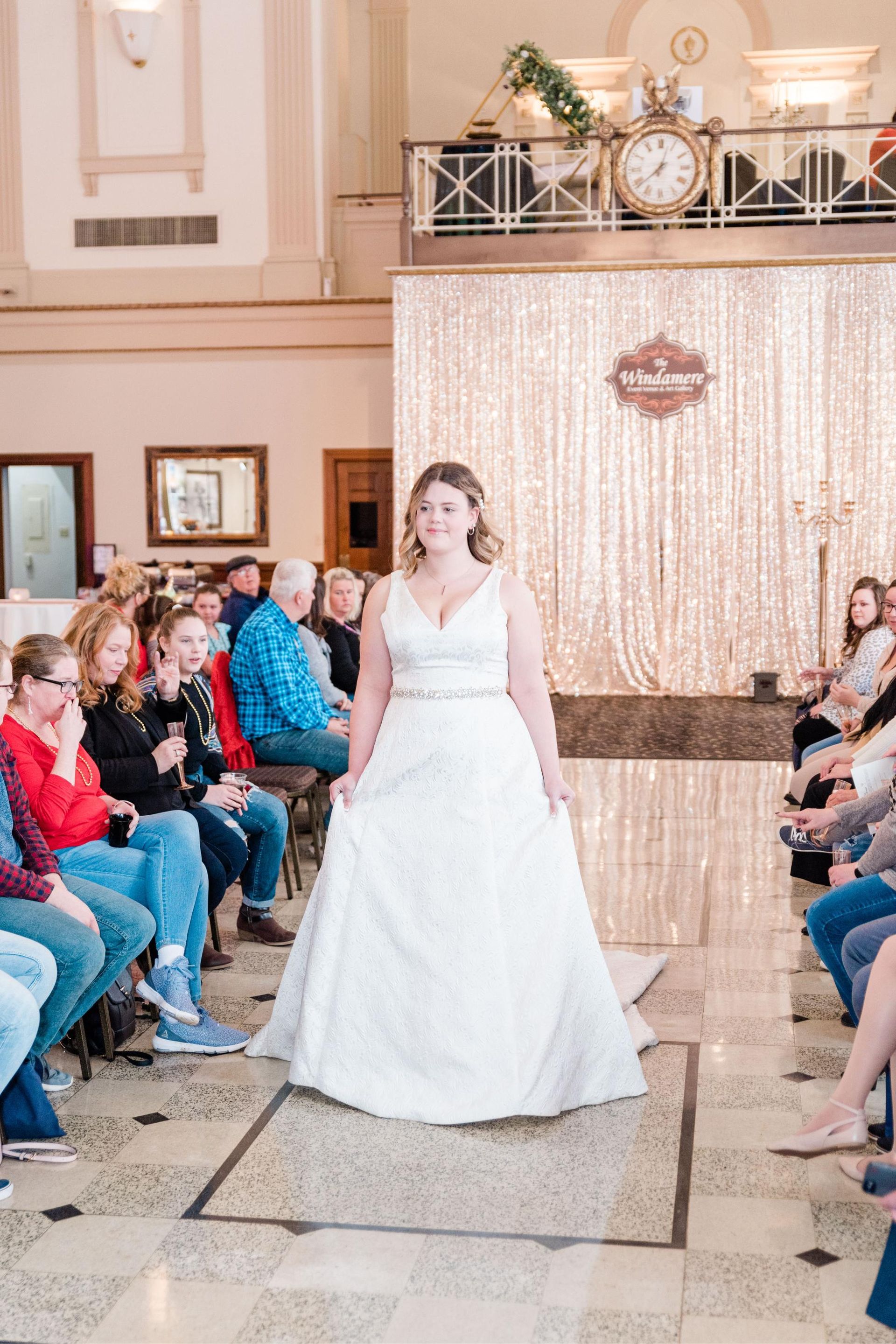 Woman in a wedding dress walks down a runway at a fashion show in a large hall.
