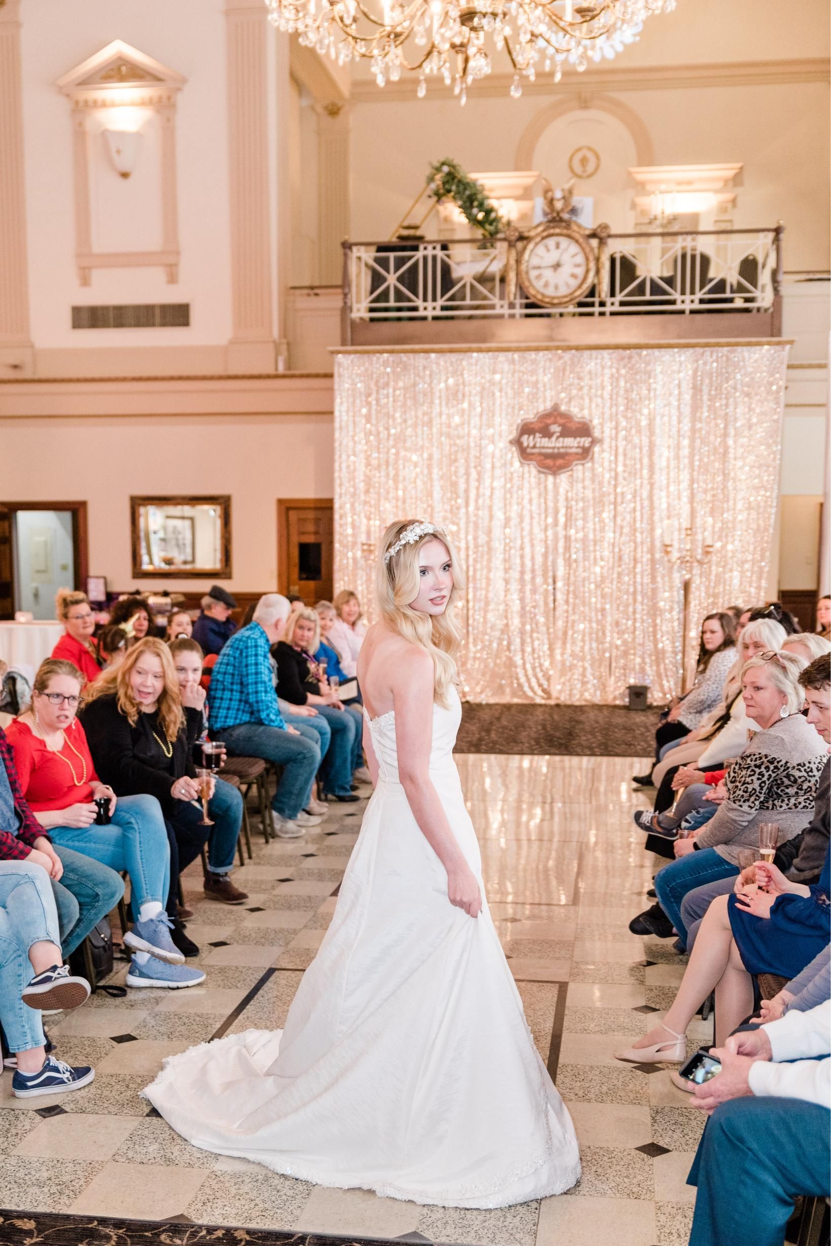 Woman in wedding dress on runway, smiling, looking over shoulder at audience. Indoors, elegant setting.