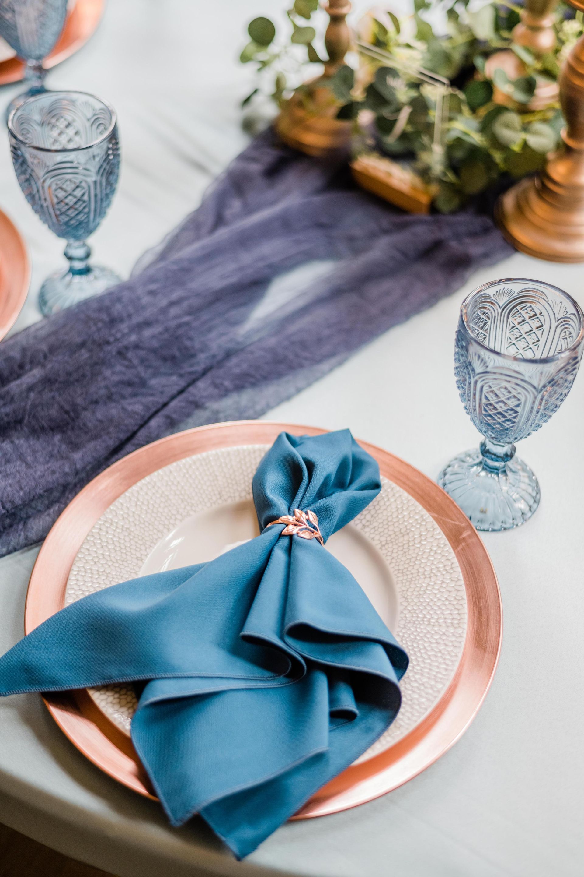 Elegant table setting: blue napkin, rose gold charger, blue goblet, floral centerpiece, blue runner.