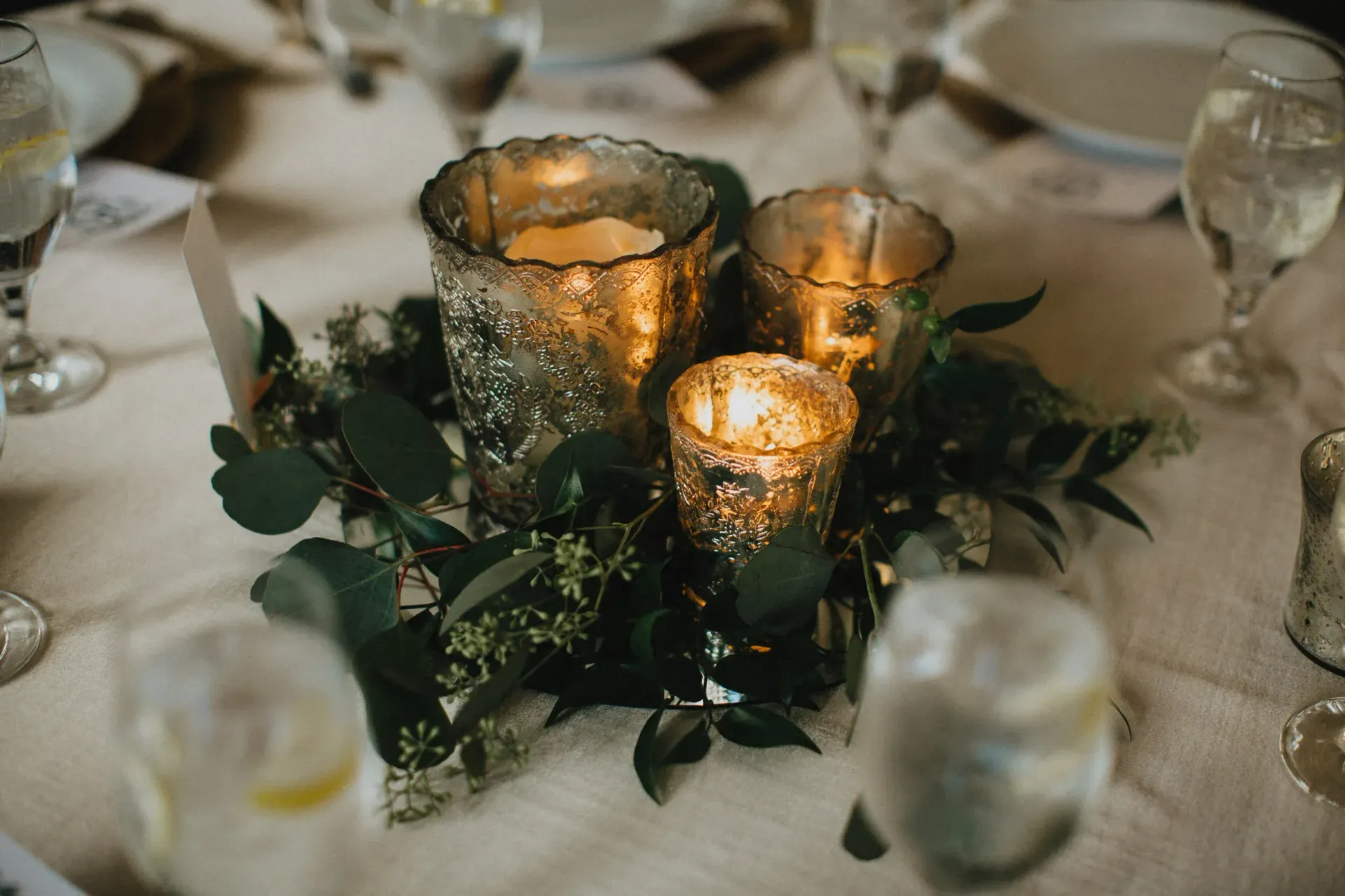 Centerpiece with three glowing gold votive candles surrounded by greenery, on a table set for a meal.