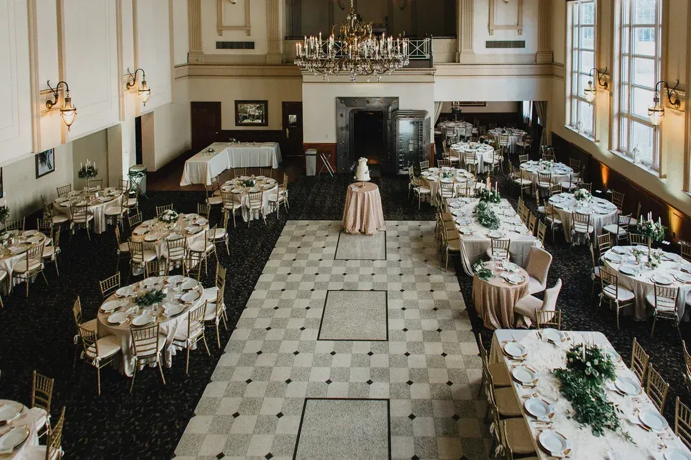 Elegant ballroom set for a wedding reception with round and rectangular tables, beige linens, and a central wedding cake.