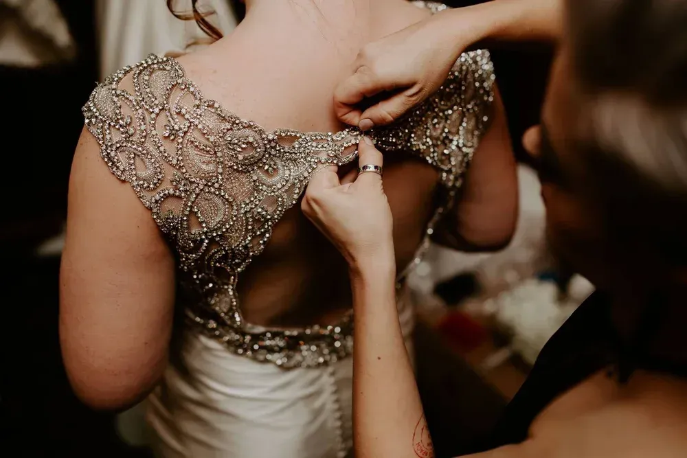 Bride in a wedding dress has the back buttoned by a person. Dress has silver beading.