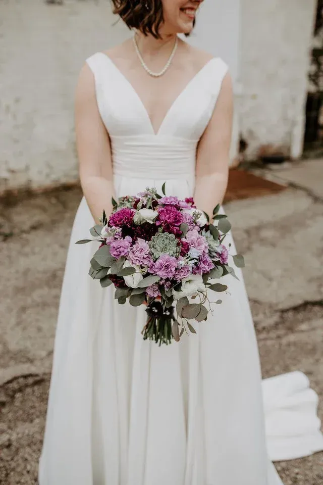 Bride in a white wedding dress holding a purple and green flower bouquet. She is smiling, and a pearl necklace is visible.