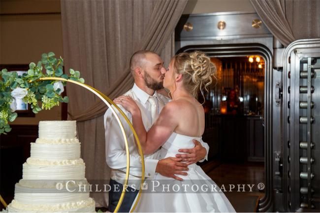 Bride and groom kiss near cake and vault door. She wears a white dress, he a white shirt.