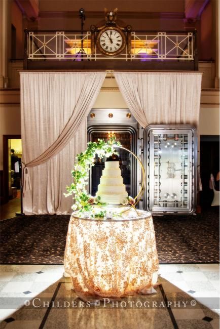 Wedding cake on gold-draped table, arch of flowers, set before an antique vault inside a grand hall.