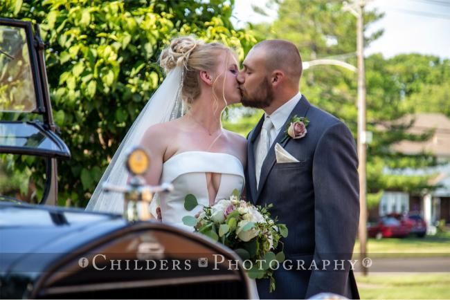Bride and groom kiss next to a vintage car. The bride wears a strapless dress and veil. The groom is in a suit.