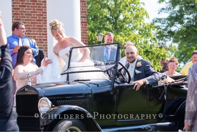Bride and groom exiting a black antique car after wedding ceremony; smiles, sunny outdoors.