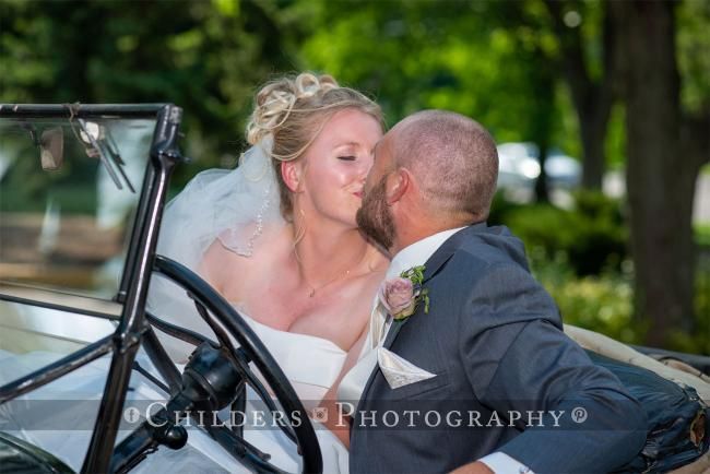 Bride and groom kiss in a vintage car. The bride wears a white dress, and the groom, a gray suit.