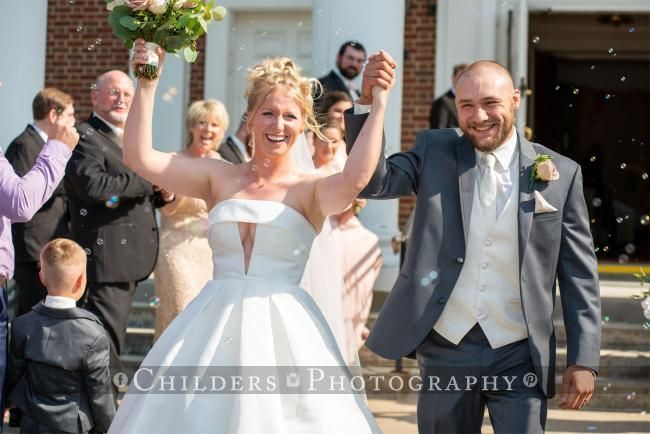 Newlyweds exiting a church, bride holding bouquet with arms raised, groom smiling, guests cheering.
