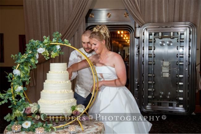 Bride and groom cutting wedding cake; gold hoop with floral arrangement, bank vault background.
