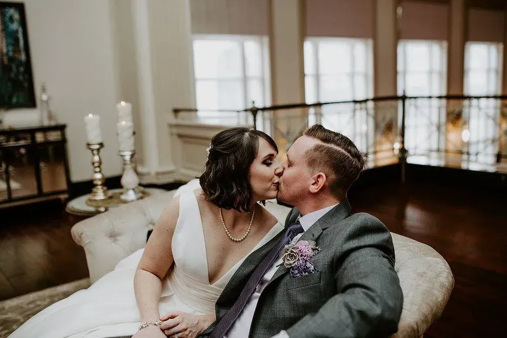 Newlyweds kissing on a cream-colored chaise lounge in a well-lit room with large windows and candles.