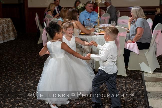 Three children dancing at a wedding reception: two girls in white dresses, and a boy in a white shirt.