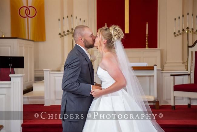 Newlyweds share a kiss at the altar in a church. Bride in white dress and veil, groom in a gray suit.