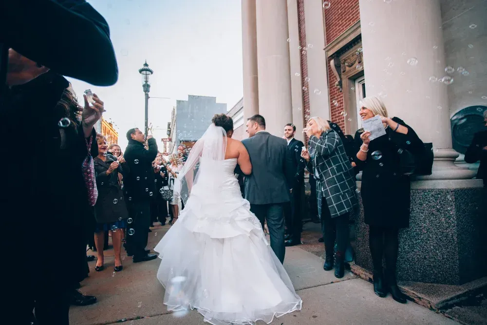 Bride and groom exit a building, walking toward the street as guests blow bubbles. 