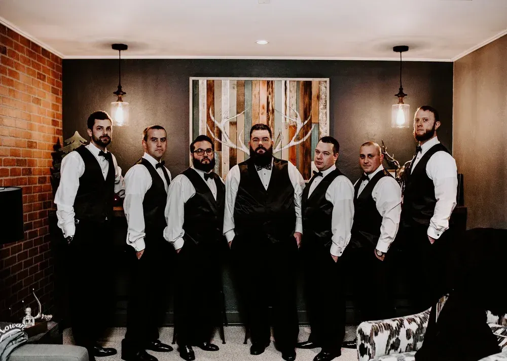 Seven men in formal attire pose for a photo. They wear black vests, white shirts,
