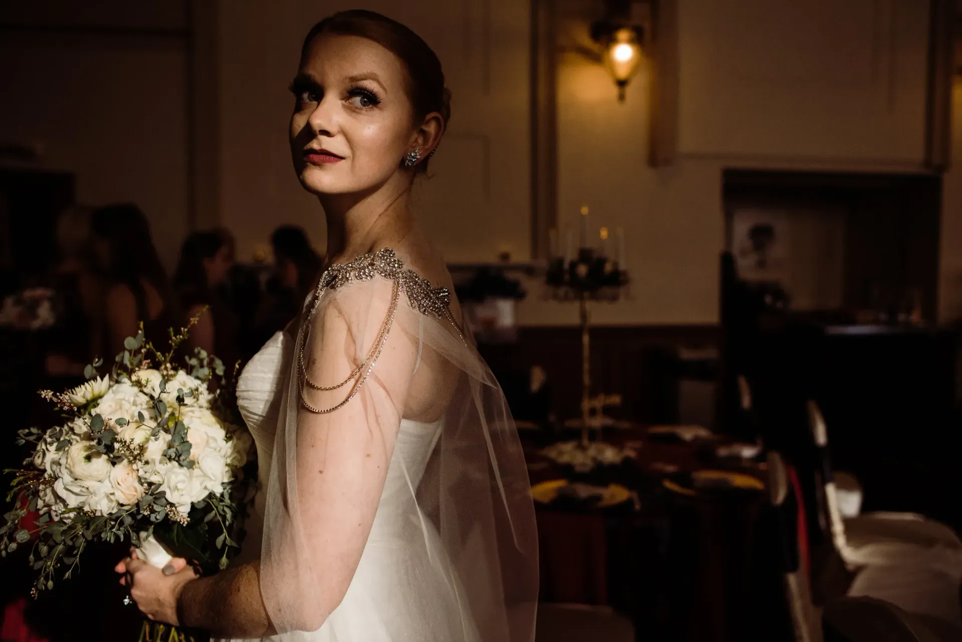 Bride in a white dress with beaded shoulder cape, holding a bouquet, looking over her shoulder in a dimly lit room.