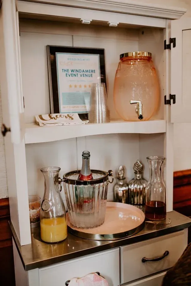 A beverage station with champagne, juice, and water dispenser in a white cabinet. A sign and glasses are on a shelf.