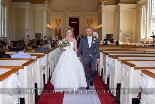 Bride and groom walk down aisle after wedding ceremony. Church interior, red carpet, white dresses, and gray suit.