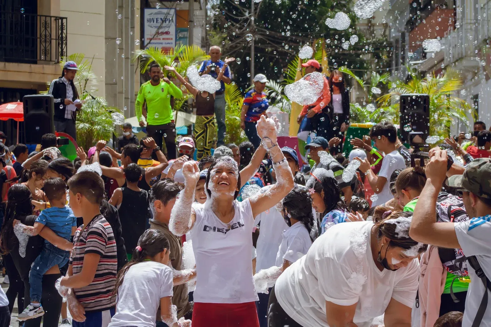 People celebrate in a crowded street, covered in foam. One woman raises her arms, enjoying the festivities.